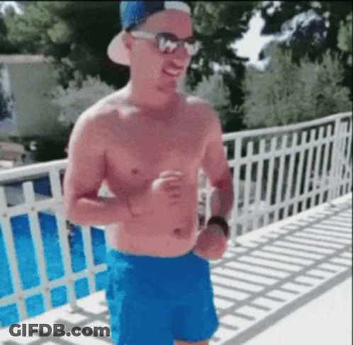 A Shirtless Man Wearing Blue Shorts And A Hat Is Standing On A Balcony . GIF