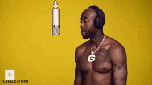 A Shirtless Man Wearing Headphones And A Necklace With A G On It Is Standing In Front Of A Microphone . GIF