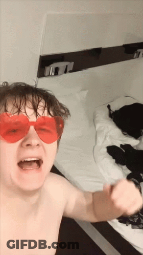 A Shirtless Man Wearing Red Heart Shaped Sunglasses Is Standing In Front Of A Bed GIF