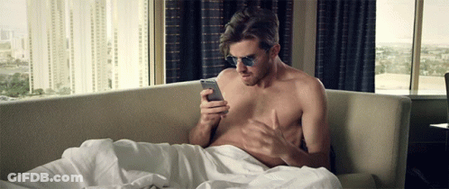 A Shirtless Man Wearing Sunglasses Is Laying On A Couch Looking At His Phone GIF