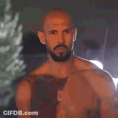 A Shirtless Man With A Beard And A Tattoo On His Chest Is Standing In Front Of A Building . GIF