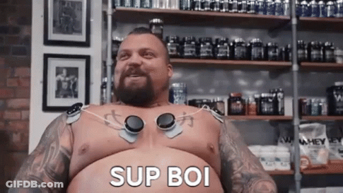 A Shirtless Man With A Beard And Tattoos Is Sitting In Front Of A Shelf With Bottles Of Supplements . GIF