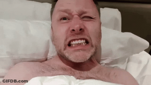 A Shirtless Man With A Beard Is Making A Funny Face While Laying In Bed GIF