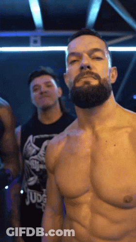 A Shirtless Man With A Beard Is Standing In A Dark Room With Other Men . GIF