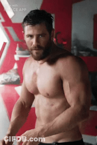 A Shirtless Man With A Beard Is Standing In Front Of A Red Wall In A Room . GIF
