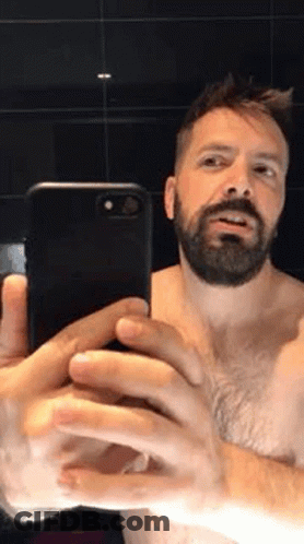 A Shirtless Man With A Beard Is Taking A Selfie With His Cell Phone In A Bathroom . GIF