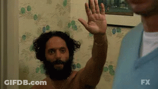 A Shirtless Man With A Beard Is Waving His Hand In Front Of A Wall . GIF