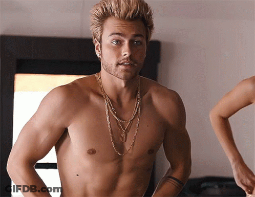 A Shirtless Man With A Necklace Around His Neck Stands In Front Of A Mirror GIF