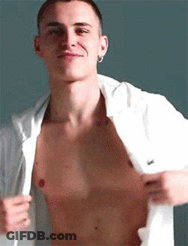 A Shirtless Man With A Tattoo On His Chest Is Taking Off His Shirt And Smiling . GIF