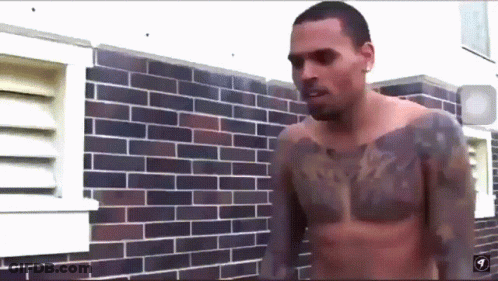 A Shirtless Man With A Tattoo On His Chest Stands In Front Of A Brick Wall GIF
