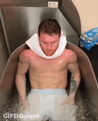 A Shirtless Man With A Towel Around His Neck Is Sitting In A Tub Of Water . GIF
