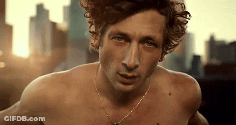 A Shirtless Man With Curly Hair Wearing A Necklace Looks At The Camera GIF