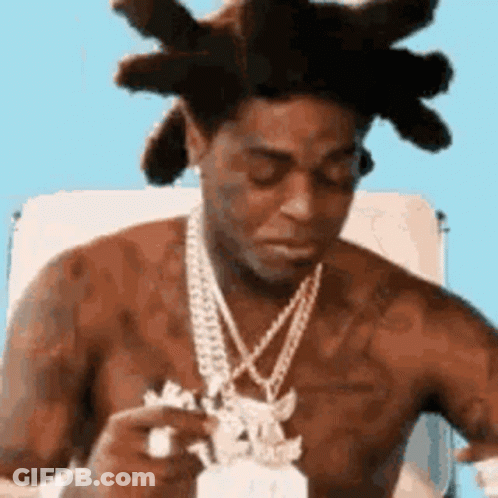 A Shirtless Man With Dreadlocks And A Necklace Is Sitting In A Chair Eating A Piece Of Food . GIF