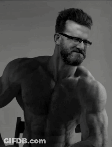 A Shirtless Man With Glasses And A Beard Is Sitting In A Chair GIF