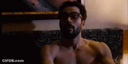 A Shirtless Man With Glasses And A Beard Is Sitting On A Couch GIF