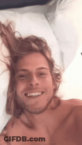 Shirtless Man With Long Hair Smiling In Bed GIF