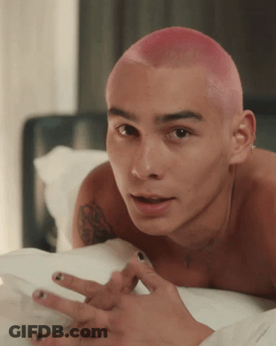 A Shirtless Man With Pink Hair And Black Nail Polish Is Laying On A Bed GIF
