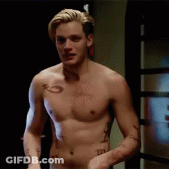 A Shirtless Man With Tattoos On His Arm And Chest Is Standing In Front Of A Door . GIF