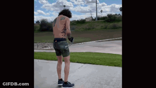 A Shirtless Man With Tattoos On His Back Is Standing On A Sidewalk . GIF