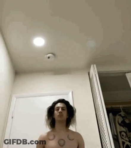 A Shirtless Man With Tattoos On His Chest Is Standing In Front Of A Door In A Bathroom . GIF