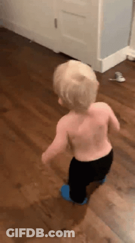 A Shirtless Toddler Is Walking On A Wooden Floor . GIF