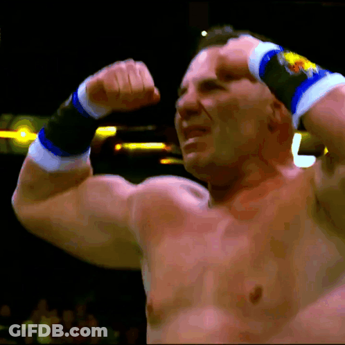 A Shirtless Wrestler Flexes His Muscles In Front Of A Crowd With The Letter L Visible In The Background GIF