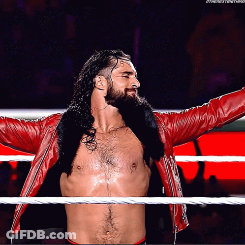 A Shirtless Wrestler In A Red Jacket Stands In A Wrestling Ring With His Arms Outstretched GIF