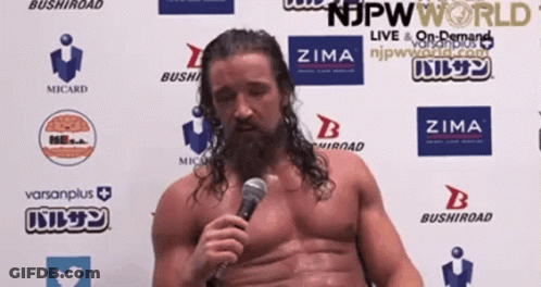 A Shirtless Wrestler Is Speaking Into A Microphone In Front Of A Wall With Logos For Njpw World And Zima GIF
