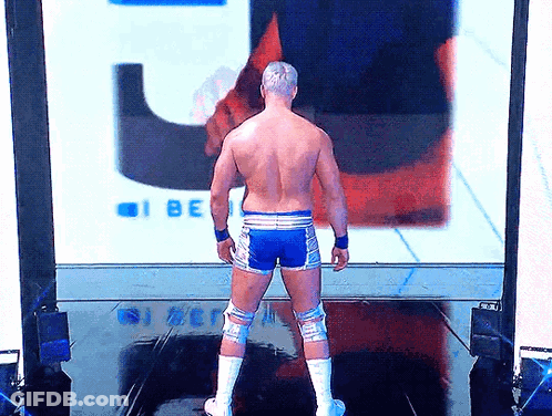 A Shirtless Wrestler Is Standing In Front Of A Large Screen That Says I Be GIF