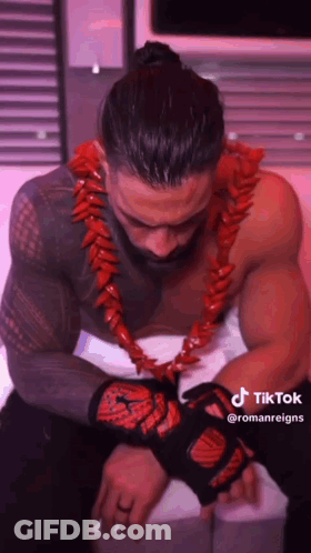 A Shirtless Wrestler Wearing A Red Lei And Gloves Is Sitting On A Bed . GIF