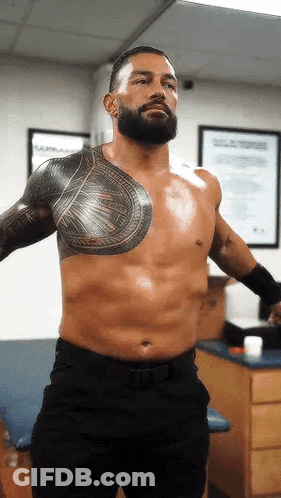 A Shirtless Wrestler With A Beard And Tattoos On His Chest Is Standing In A Room . GIF