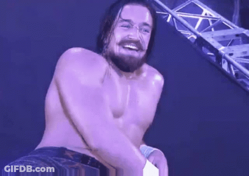 A Shirtless Wrestler With A Beard Is Smiling While Standing In Front Of A Purple Wall . GIF