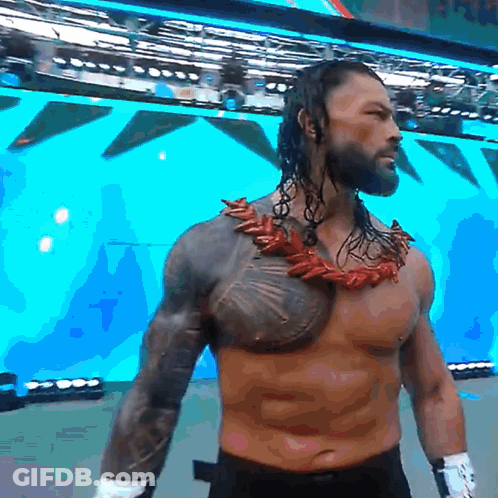 A Shirtless Wrestler With A Necklace Around His Neck Stands In Front Of A Large Screen GIF