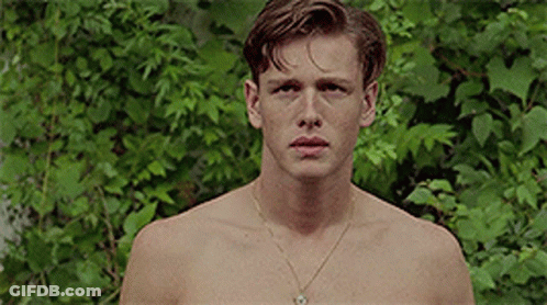 A Shirtless Young Man Wearing A Gold Necklace Is Standing In Front Of A Bush . GIF