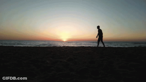 A Silhouette Of A Man Walking On A Beach At Sunset GIF