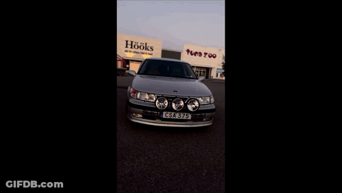 A Silver Car Is Parked In Front Of A Building That Says Toys Too GIF