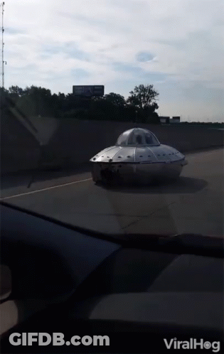 A Silver Flying Saucer Is Driving Down A Highway With A Billboard In The Background GIF