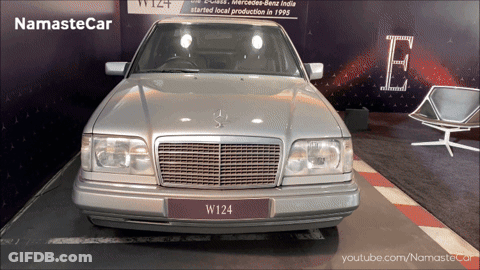A Silver Mercedes W124 Is On Display In Front Of A Sign That Says Namaste Car GIF