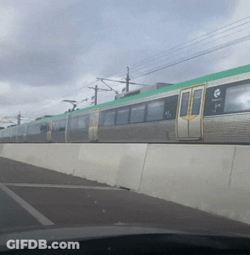A Silver Train With A Green Top Is Going Down A Road GIF