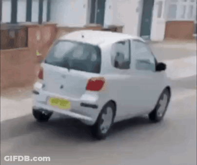 A Small White Car Is Driving Down A Street Next To A House . GIF