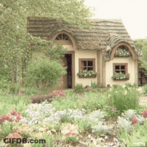 A Small White House With A Thatched Roof Is Surrounded By Flowers And Trees . GIF