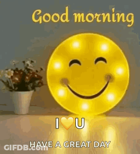 A Smiley Face Is Sitting On A Table Next To A Vase Of Flowers And A Good Morning Message . GIF