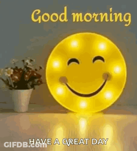 A Smiley Face Is Sitting On A Table Next To A Vase Of Flowers And Says `` Good Morning Have A Great Day '' . GIF