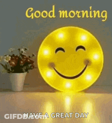 A Smiley Face Is Sitting On A Table Next To A Vase Of Flowers And Says `` Good Morning Have A Great Day '' . GIF