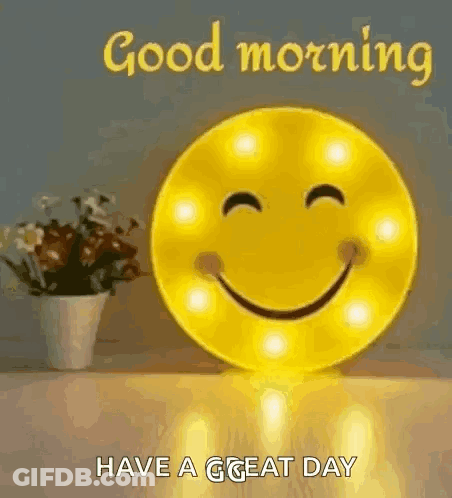 A Smiley Face Is Sitting On A Table Next To A Vase Of Flowers And Says `` Good Morning Have A Great Day '' . GIF