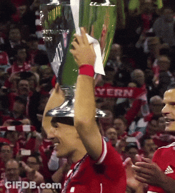 A Soccer Player Holds Up A Trophy In Front Of A Crowd With A Bayern Scarf Behind Him GIF