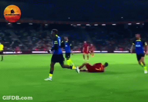 A Soccer Player In A Blue Jersey Is Running Towards A Player In A Yellow Jersey Who Is Laying On The Ground GIF