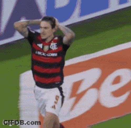 A Soccer Player In A Red And Black Jersey Stands On A Field In Front Of A Sign That Says Be GIF