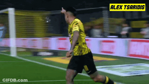 A Soccer Player In A Yellow Jersey Is Running On The Field With Alex Tsaridis In The Background GIF