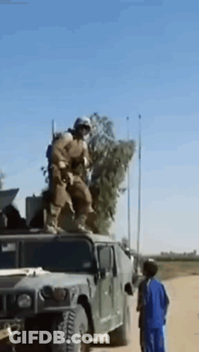 A Soldier Is Standing On Top Of A Military Vehicle While A Boy Looks On . GIF